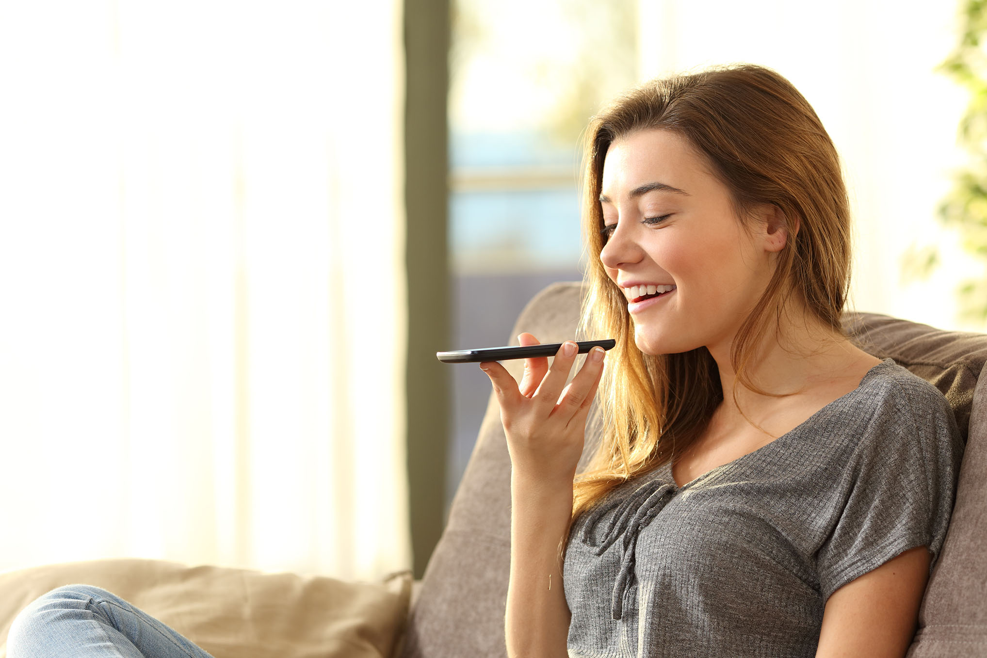 Voss Give Teams a Voice image - Girl using a smart phone voice recognition on line sitting on a sofa in the living room at home with a warm light and a window in the background