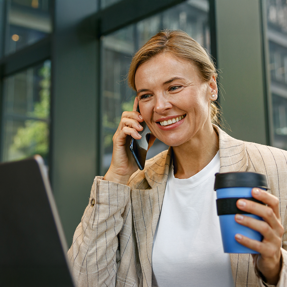 Woman on a business call on mobile phone looking at laptop holding a coffee